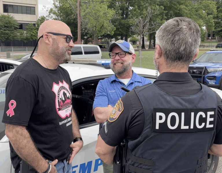 John Covington stands next to a parked Memphis Police Department vehicle, smiling and talking with two officers—one facing him and one seen from behind—during an outdoor community event.